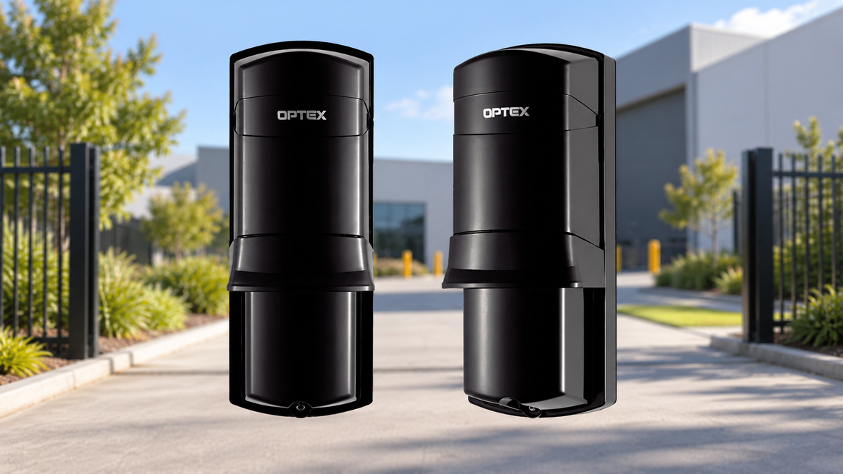 Two Optex outdoor security beam detectors with a commercial driveway and gated warehouse entrance in the background.