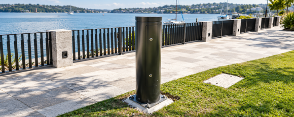 Black security bollard installed beside a waterfront promenade with marina, railing and landscaped grass in the background.