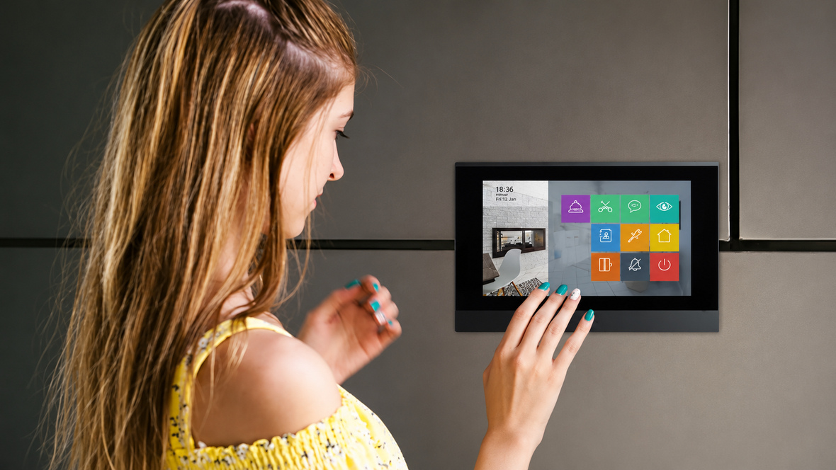 Woman using a wall-mounted indoor touchscreen intercom monitor with home control options.