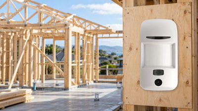 Security detector with integrated camera mounted on a plywood panel beside exposed timber framing at a residential construction site.